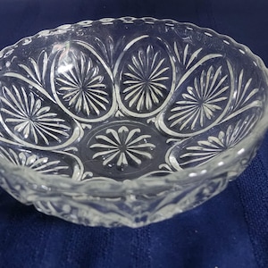 May include: A clear glass bowl featuring a pressed pattern of radiating sunburst or starburst motifs within oval panels around the interior sides and a central circular starburst design at the base, finished with a scalloped, textured rim.