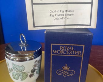 May include: A Royal Worcester egg coddler with a silver lid and handle, featuring a white ceramic body decorated with green leaves and blackberries. The box and recipe card are also visible, with the Royal Worcester logo and text.