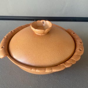 May include: A tan ceramic casserole dish with a lid. The lid has a small, flower-shaped handle. The dish has a decorative, raised border. The casserole dish is oval-shaped and appears to be vintage.
