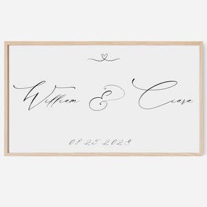 Könnte beinhalten: Gerahmter Druck mit hellem Holzrahmen. Der Druck zeigt die Namen "William & Ciara" in eleganter schwarzer Schrift, mit einem kleinen Herzen über den Namen und dem Datum "08.25.2023" darunter. Der Hintergrund ist weiß.