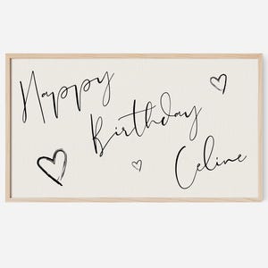 Pode incluir: Uma placa de lona bege com moldura de madeira. A placa tem texto escrito à mão em preto que diz "Happy Birthday Celine" com três corações pequenos.