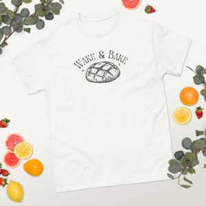 Può includere: T-shirt bianco con un'illustrazione in bianco e nero di un pane e la scritta "WAKE & BAKE". La maglietta è circondata da frutta fresca, tra cui fragole, arance e limoni.