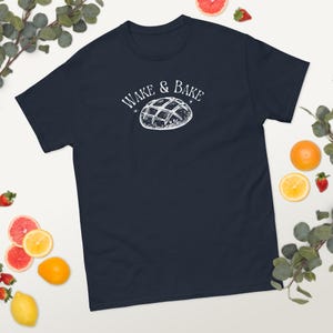 Può includere: T-shirt blu navy con la scritta "WAKE & BAKE" sopra un'illustrazione bianca di un waffle. La maglietta è circondata da frutta e verde.