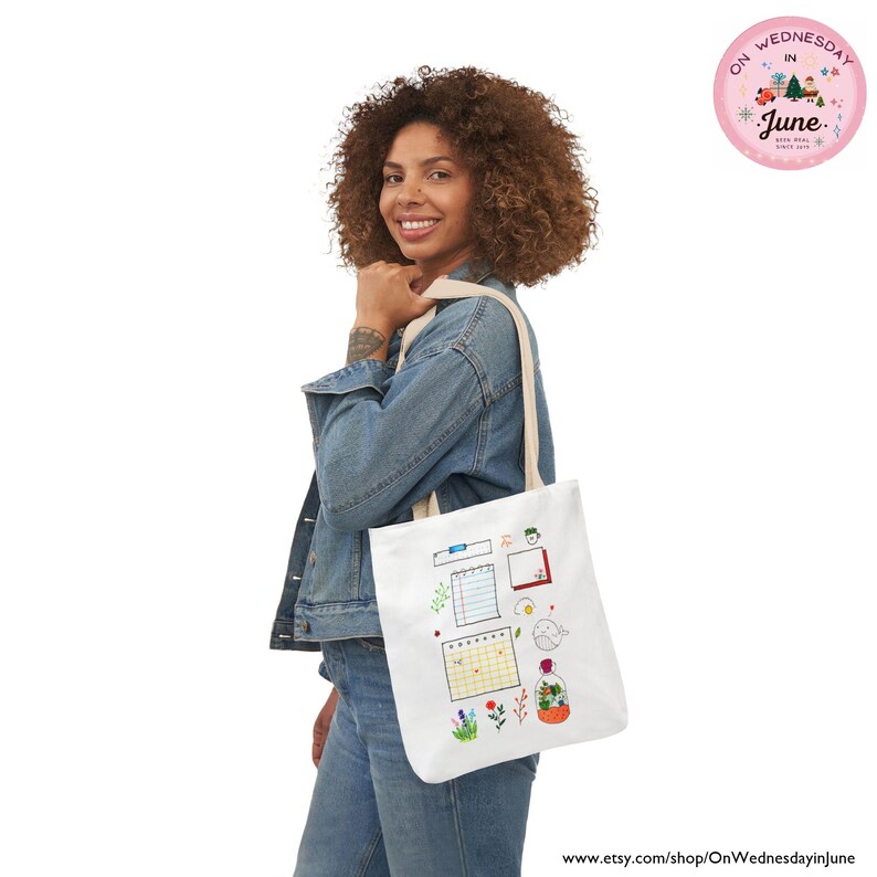 Puede incluir: Un bolso tote blanco con una colorida ilustraci&oacute;n de un calendario, notas y flores. El bolso lo lleva una persona que lleva una chaqueta vaquera. Una pegatina circular rosa con texto est&aacute; en la esquina superior derecha.
