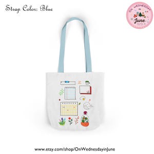 Puede incluir: Bolso tote blanco con correas azul claro. La bolsa presenta un dise&ntilde;o caprichoso con elementos dibujados a mano que incluyen un calendario, un cuaderno, una ballena y flores. El texto "Strap Color: Blue" est&aacute; en la parte superior izquierda.