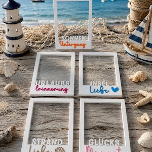 Könnte beinhalten: Fünf weiße, rechteckige Fotorahmen mit ausgeschnittenen Mitten. Jeder Rahmen trägt erhabene Schriftzüge: SONNEN Untergang, URLAUBS Erinnerung, INSEL Liebe mit blauem Herz, STRAND Liebe mit Muschel-Symbol sowie GLUCKS Moment in verschiedenen Farben.