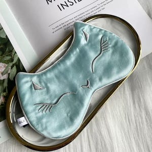 May include: A light blue silk sleep mask with embroidered facial features, including closed eyes and a nose. The mask is on a gold-rimmed tray with a white interior. The word "SILK" is visible on the mask.