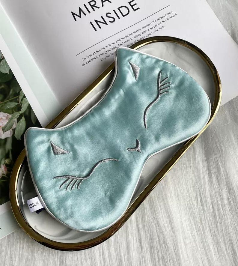 May include: A light blue silk sleep mask with embroidered facial features, including closed eyes and a nose. The mask is on a gold-rimmed tray with a white interior. The word "SILK" is visible on the mask.