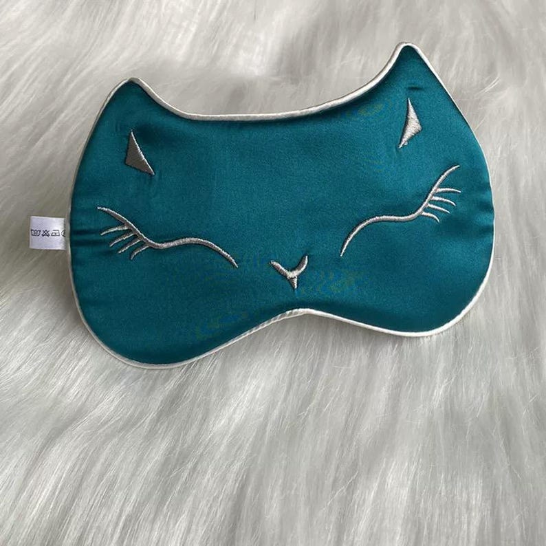 May include: Teal-colored sleep mask with a cat face design. The mask features embroidered silver eyelashes, nose, and triangular accents. It has a white trim and is made of a soft, silky material. The mask is designed to block out light for better sleep.