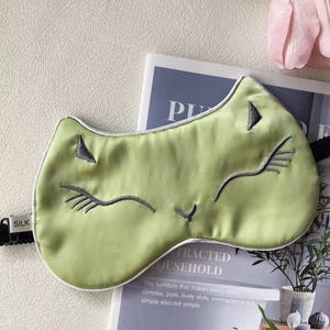 May include: A light green silk sleep mask with a cat face design. The mask features embroidered gray details for the eyes, nose, and whiskers, and a white trim. The black elastic band has "SILK" printed on it.