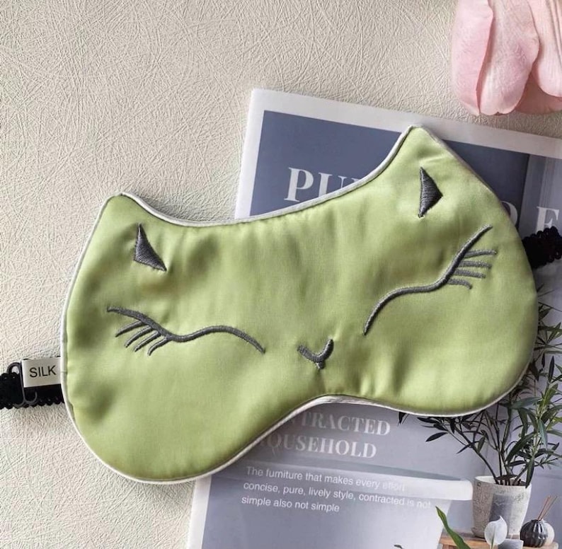 May include: A light green silk sleep mask with a cat face design. The mask features embroidered gray details for the eyes, nose, and whiskers, and a white trim. The black elastic band has "SILK" printed on it.