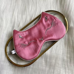 May include: A pink silk sleep mask with embroidered cat face details, including closed eyes and a nose. The mask is on a clear tray with a gold rim. A small tag reads "SILK".