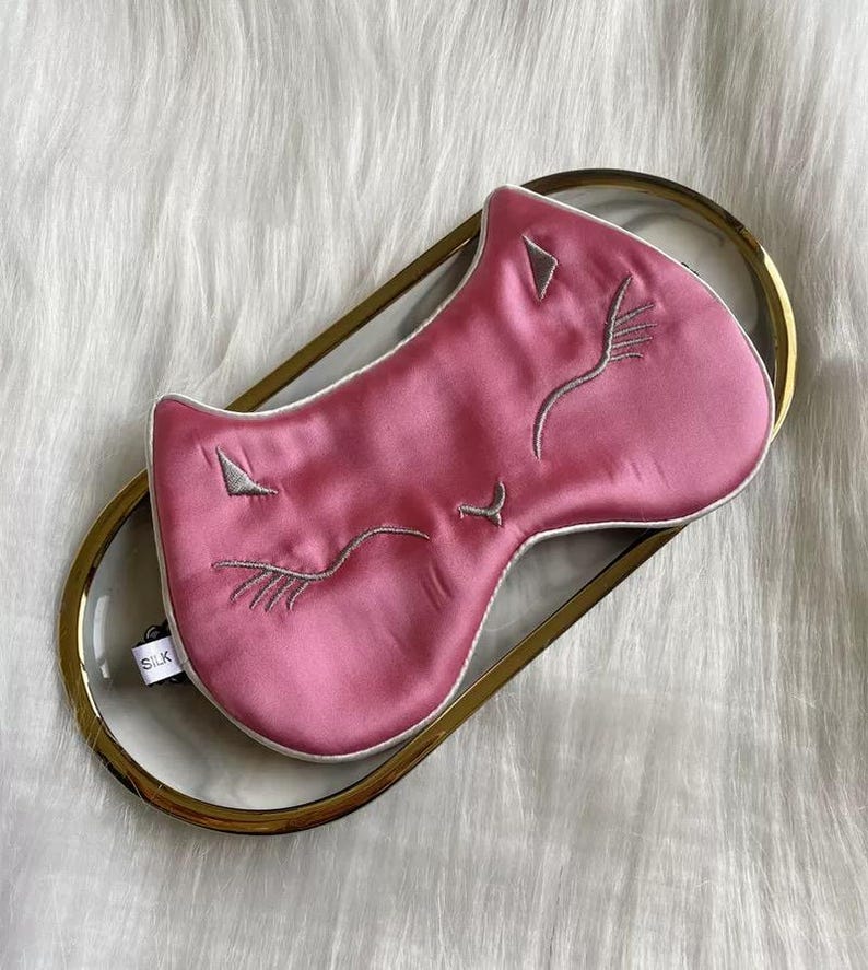 May include: A pink silk sleep mask with embroidered cat face details, including closed eyes and a nose. The mask is on a clear tray with a gold rim. A small tag reads "SILK".
