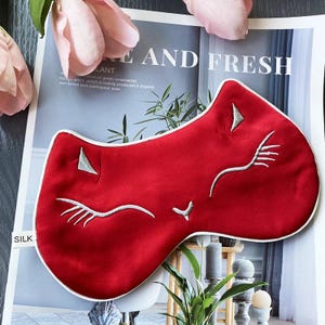 May include: A red silk sleep mask in the shape of a cat's face, with white embroidered details and a white border. The mask has a smooth texture and is designed to cover the eyes. The word "SILK" is visible on the left side of the image.