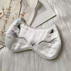May include: A silver-colored sleep mask shaped like a cat's face, with embroidered closed eyes, eyelashes, and nose. The mask is made of a soft, silky material. The background includes a white, fluffy surface and a piece of clothing with floral embroidery.