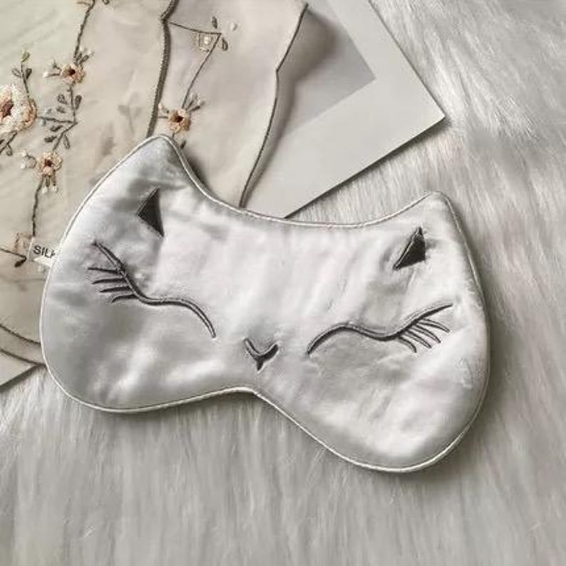 May include: A silver-colored sleep mask shaped like a cat's face, with embroidered closed eyes, eyelashes, and nose. The mask is made of a soft, silky material. The background includes a white, fluffy surface and a piece of clothing with floral embroidery.