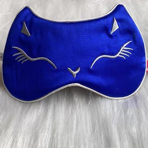 May include: A blue satin sleep mask shaped like a cat's face, with embroidered silver eyelashes, nose, and ear details. The mask has a white trim and is designed to block out light for better sleep. The mask is laying on a white, fluffy surface.