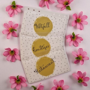 Op de afbeelding: Transparante plastic organizer met drie vakken, elk met een gouden cirkel met zwarte tekst. De tekst luidt "Notfall", "Sonstiges" en "Restaurant". De organizer is versierd met kleine zwarte stippen en omgeven door roze bloemen.