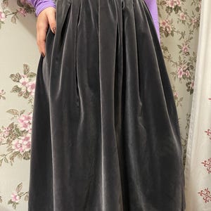 May include: A dark gray velvet skirt with a high waist and pleated design. The skirt is knee-length and has a soft, flowing appearance. The fabric has a subtle sheen, and the skirt appears to be well-constructed.