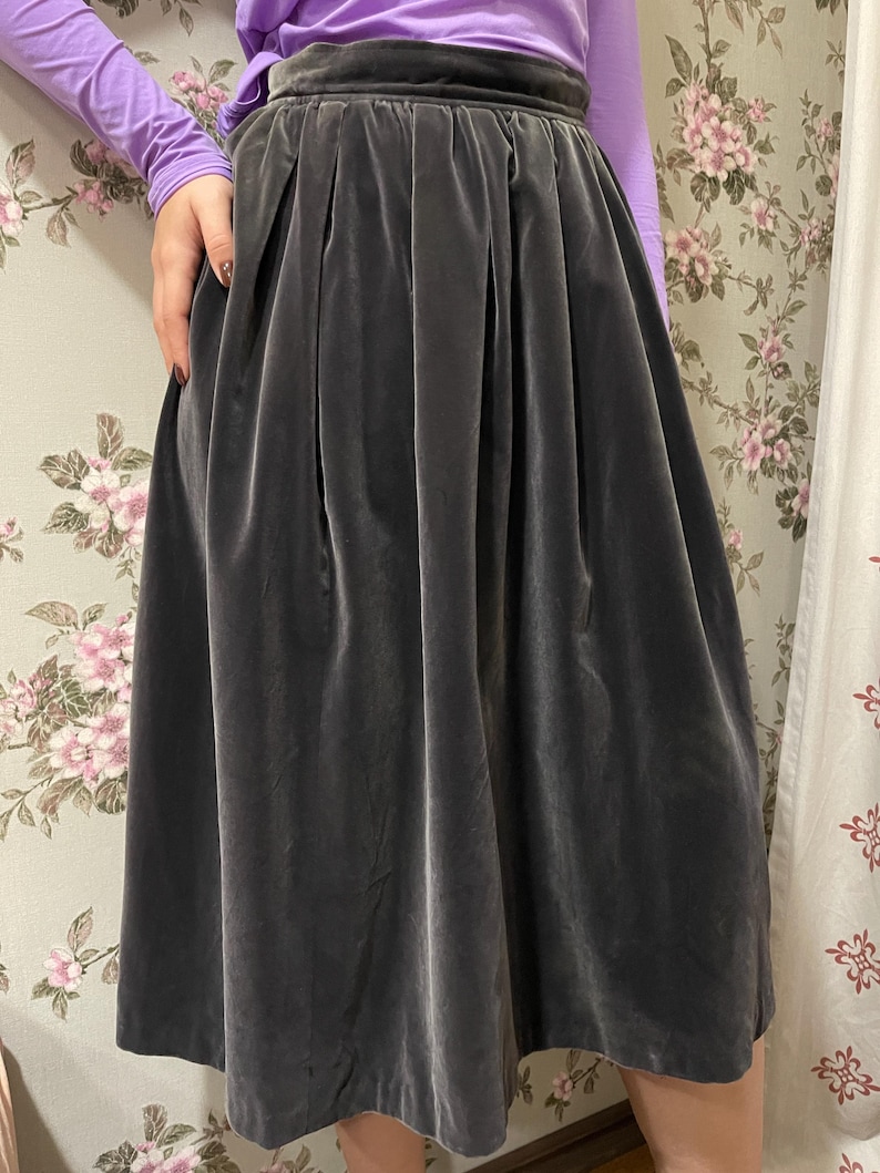 May include: A dark gray velvet skirt with a high waist and pleated design. The skirt is knee-length and has a soft, flowing appearance. The fabric has a subtle sheen, and the skirt appears to be well-constructed.