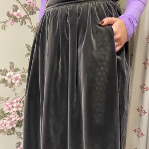 May include: A dark gray velvet skirt with a high waist and pockets. The skirt has a full, flowing silhouette and is made of a soft, luxurious fabric. The person is wearing a long-sleeved purple shirt.