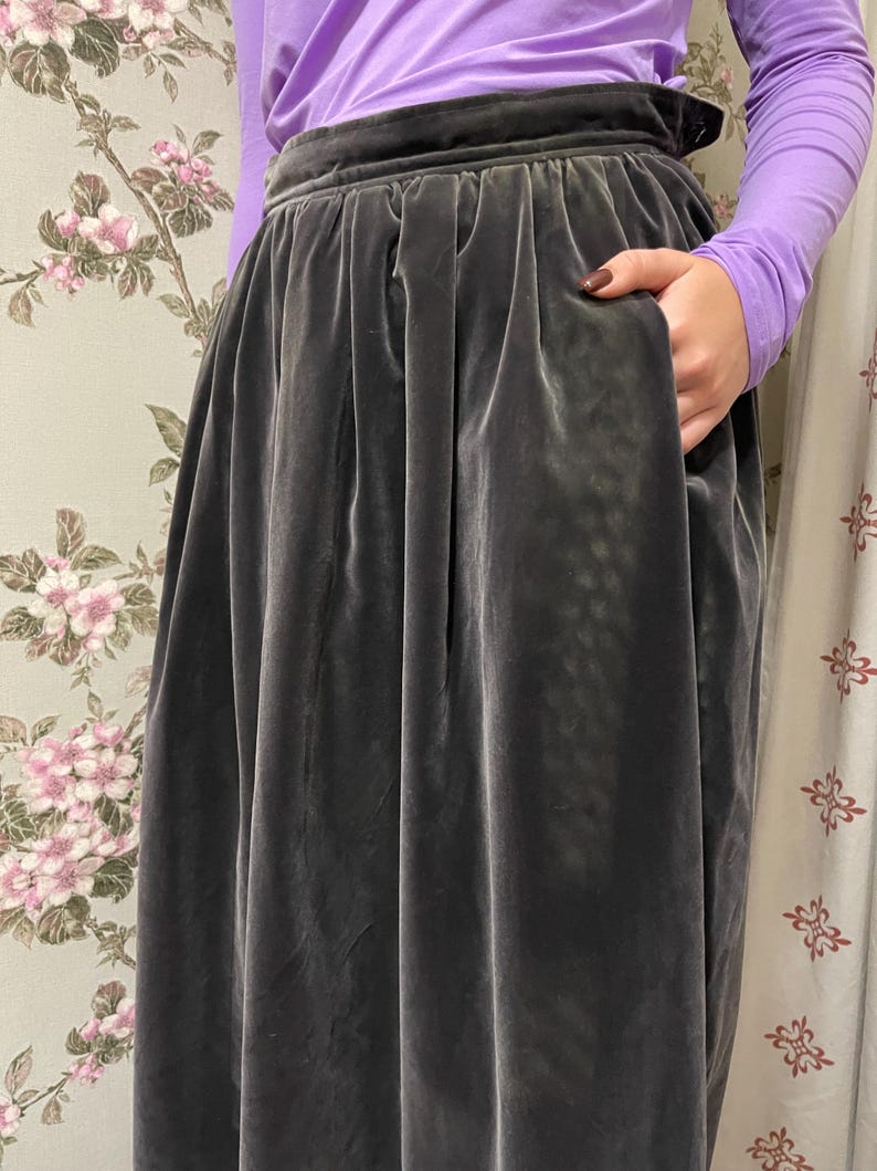 May include: A dark gray velvet skirt with a high waist and pockets. The skirt has a full, flowing silhouette and is made of a soft, luxurious fabric. The person is wearing a long-sleeved purple shirt.