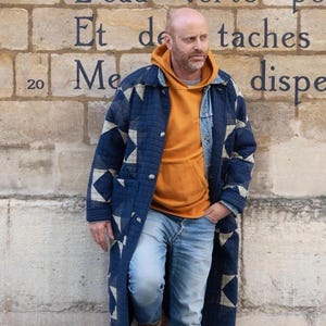 May include: A man wearing a blue quilted jacket with white geometric patterns, a bright orange hooded sweatshirt, blue jeans, and brown boots. He is standing in front of a stone wall with French text.