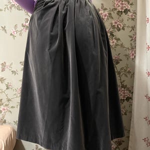 May include: A dark gray velvet skirt with a high waist and a pleated design. The skirt is knee-length and has a flowing silhouette. The fabric appears soft and drapes well. The background features a floral wallpaper.