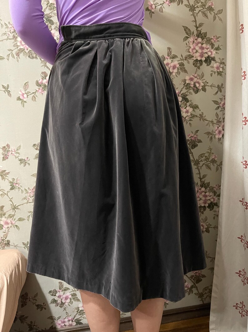 May include: A dark gray velvet skirt with a high waist and a pleated design. The skirt is knee-length and has a flowing silhouette. The fabric appears soft and drapes well. The background features a floral wallpaper.