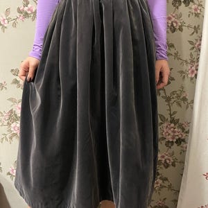 May include: A dark gray velvet skirt with a high waist and a full, pleated design. The skirt is knee-length and has a soft, flowing appearance. The model is wearing a long-sleeved purple shirt.