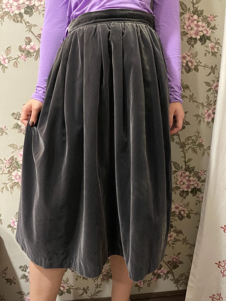May include: A dark gray velvet skirt with a high waist and a full, pleated design. The skirt is knee-length and has a soft, flowing appearance. The model is wearing a long-sleeved purple shirt.