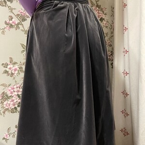 May include: A dark gray velvet skirt with a high waist and a wide waistband. The skirt has a pleated design and falls to a midi length. The skirt is paired with a long-sleeved purple top.