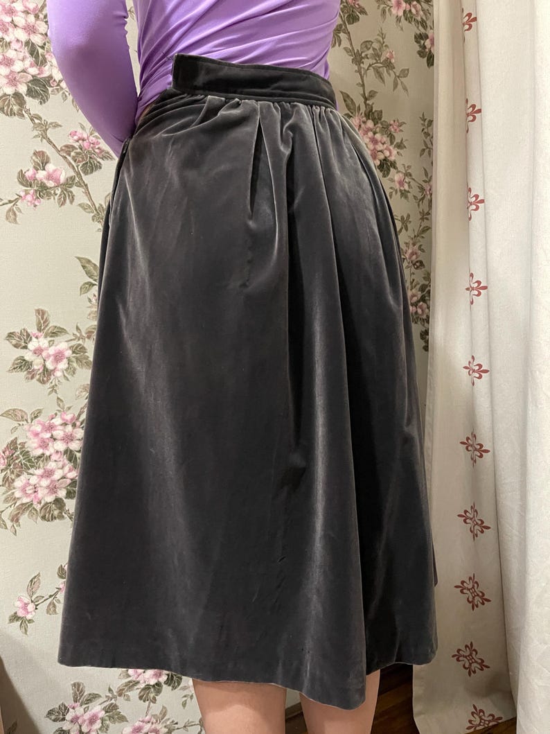 May include: A dark gray velvet skirt with a high waist and a wide waistband. The skirt has a pleated design and falls to a midi length. The skirt is paired with a long-sleeved purple top.