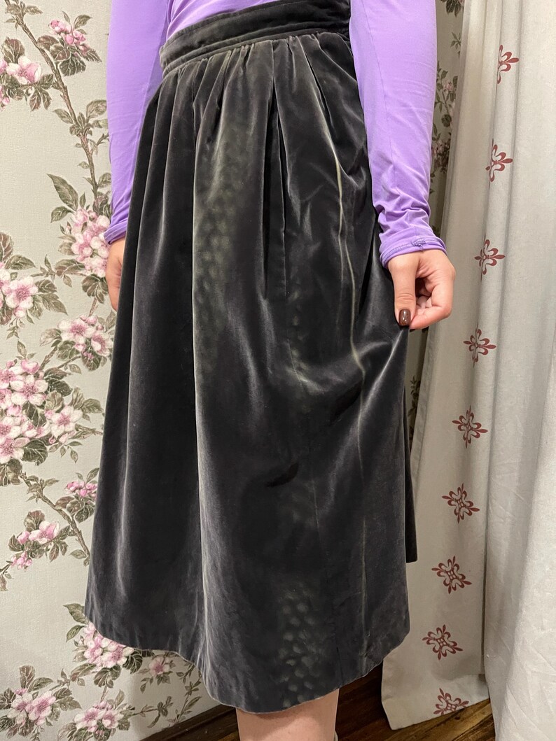 May include: A dark gray velvet skirt with a high waist and pleats. The skirt is knee-length and has a soft, flowing appearance. The skirt is paired with a long-sleeved purple top.