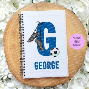 May include: A white notebook with a blue letter G and the name George printed in blue. The letter G has a pair of black football boots hanging from it and a black and white football is to the right of the letter. The notebook has a spiral binding.