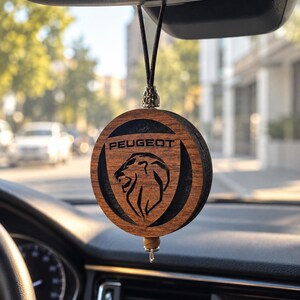 Puede incluir: Un ambientador de coche circular de madera colgado del espejo retrovisor. La parte frontal presenta un acabado de madera marrón oscuro con un logotipo de león Peugeot grabado en negro y el texto PEUGEOT. Está suspendido por un cordón negro con cuentas plateadas.