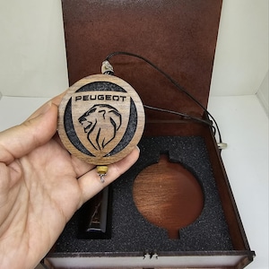 Może przedstawiać: Drewniany wisiorek z logo Peugeot z czarnym sznurkiem, prezentowany w brązowym drewnianym pudełku. Wisiorek przedstawia nazwę Peugeot i emblemat lwa. Pudełko ma ciemne wnętrze z wycięciem na wisiorek i mały przedmiot.
