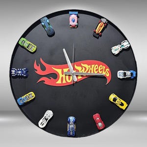 May include: Black round clock featuring miniature Hot Wheels cars as hour markers. The clock face has the Hot Wheels logo in red and yellow. The clock hands are silver.