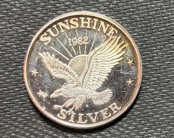 May include: A silver round coin with the words "Sunshine Silver 1982" around the edge. A bald eagle with its wings spread is in the center of the coin.