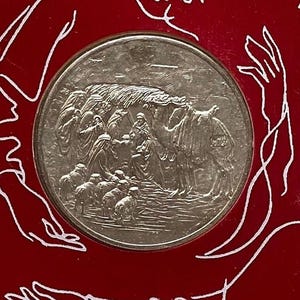 May include: A silver coin with a raised design depicting a scene from the Bible. The scene shows a group of people with a camel and sheep. The coin is surrounded by a red background.