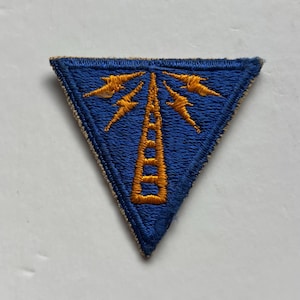 May include: A triangular embroidered patch featuring a blue background with a central yellow tower graphic. The tower is flanked by two yellow lightning bolt symbols on each side. The patch has a thick blue stitched border and a tan fabric backing.