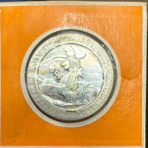 May include: A silver-colored Winners Inn casino token. The token features an illustration of a cartoon character wearing a hat and holding a drink. The token is set in a square orange and white cardboard frame.