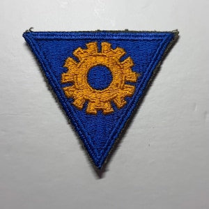 May include: A triangular embroidered patch featuring a vibrant blue background with a centered golden-yellow gear icon. The edges of the patch are finished with a dark olive-green stitched border, creating a distinct contrast against the blue fabric.