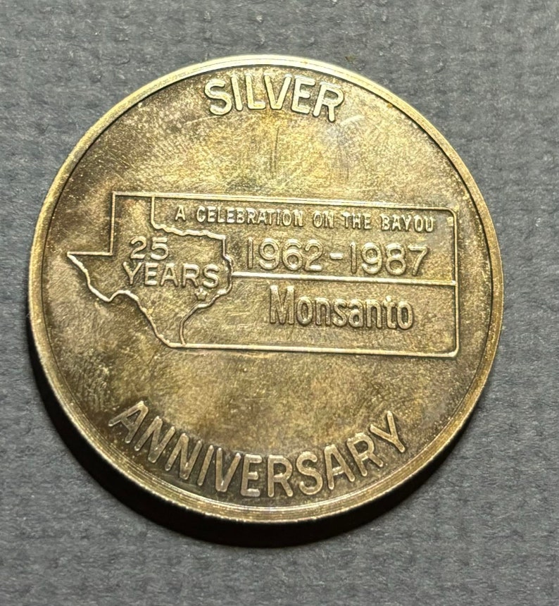 May include: A silver commemorative coin celebrating the 25th anniversary of Monsanto in 1987. The coin features a map of the state of Missouri with the text "A Celebration on the Bayou 1962-1987 Monsanto 25 Years Anniversary".