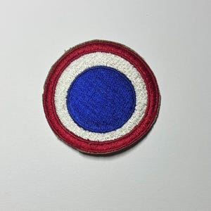 May include: A circular embroidered patch featuring a concentric design with a solid blue center, a white middle ring, and a red outer border. The stitching is dense and textured, creating a raised surface with a defined edge on a plain background.