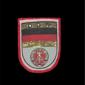 May include: A rectangular embroidered patch with a red border featuring the letters D.D.R. in gold at the top. The center displays horizontal black, red, and gold stripes above a grey field containing a circular emblem with a hammer and compass.