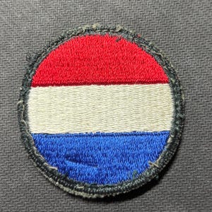May include: A circular embroidered patch featuring a horizontal tricolor design of red, white, and blue stripes. The patch has a dark, stitched border that frames the vibrant fabric colors, creating a clean and defined edge for this textile emblem.