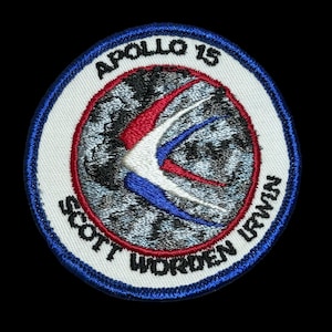 May include: A white embroidered patch with a blue border. The patch features a stylized image of a white bird with red and blue accents flying over a gray and white image of the Earth. The text "APOLLO 15" is printed in black at the top of the patch, and the text "SCOTT WORDEN IRVIN" is printed in black around the bottom of the patch.