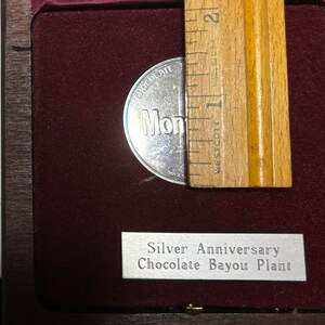 May include: A silver coin with the words "Chocolate Bayou Plant" and "Mondelez" on it. The coin is in a wooden box with a silver plaque that reads "Silver Anniversary Chocolate Bayou Plant".