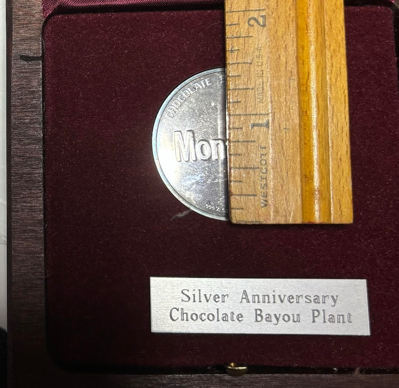May include: A silver coin with the words "Chocolate Bayou Plant" and "Mondelez" on it. The coin is in a wooden box with a silver plaque that reads "Silver Anniversary Chocolate Bayou Plant".